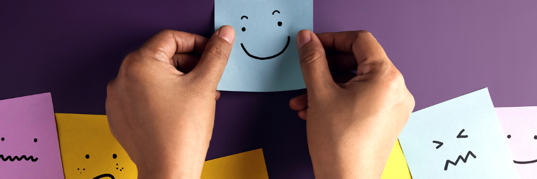 TPerson sticking a post it with a smile drawn on it onto a purple wall with other post it faces surrounding it