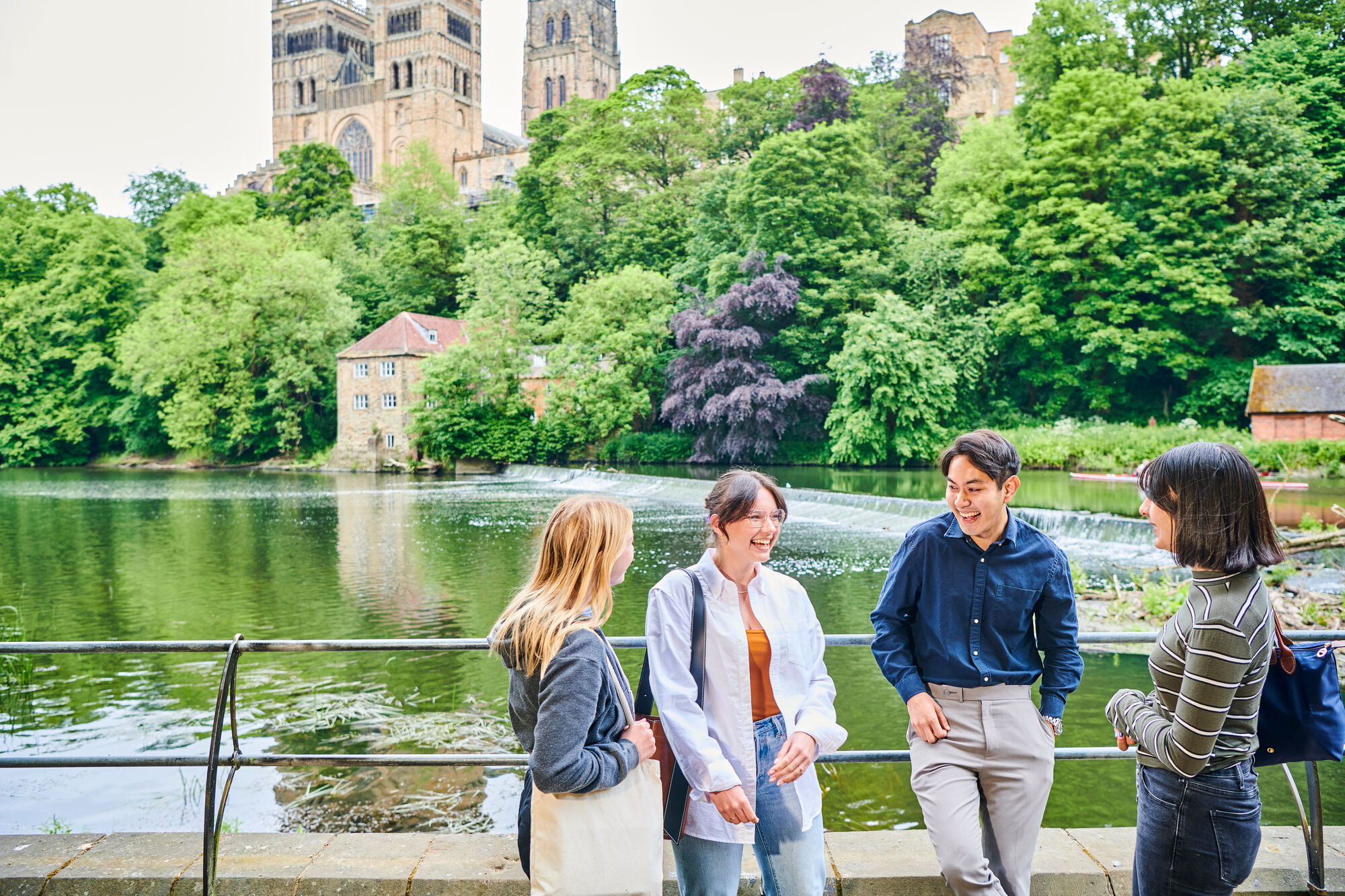 A group of students chatting on a bridge in front of a river, with lots of greenery and Durham Cathedral in the background