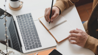 TClose up of a persons hands taking notes in a notebook in front of their laptop