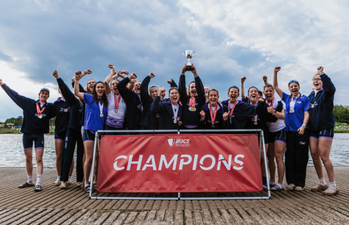 A group of mainly women standing on the banks of a river celebrating while holding a trophy and standing in front of a red BUCS champions banner