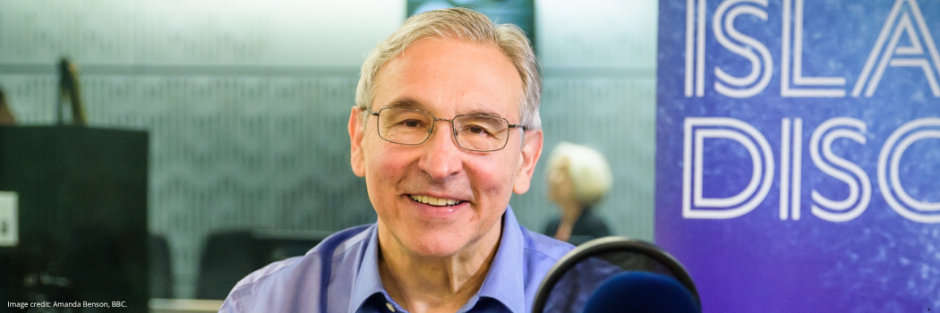 Professor Carlos Frenk smiling into the camera with a the a radio microphone in the foreground and the edge of some blue branding behind him