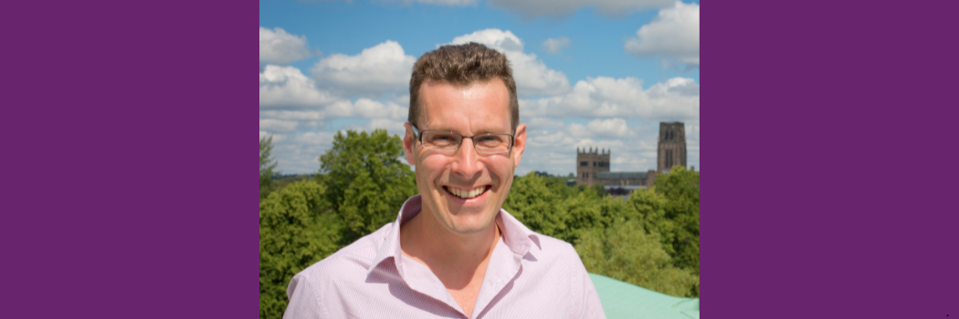 Professor Simon Cornish standing in the sunshine with a view of treetops and Durham Cathedral behind him