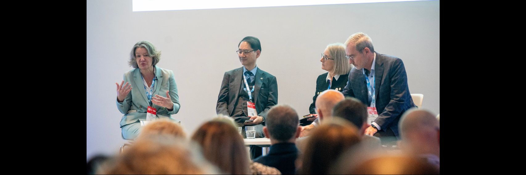 Four people sitting at a desk chatting, in front of an audience seen from behind