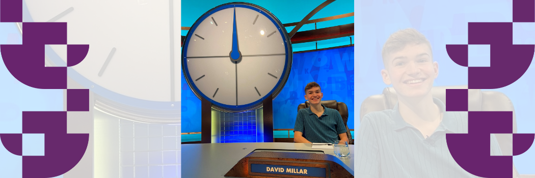 A smiling young man with his name lit up sits at a desk near a large clock