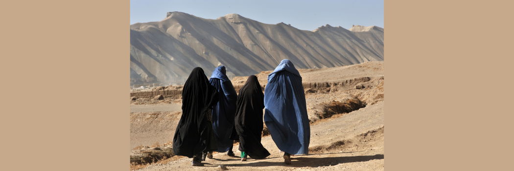 Four Afghan women in blue and black burqa walking on dirt path in barren land