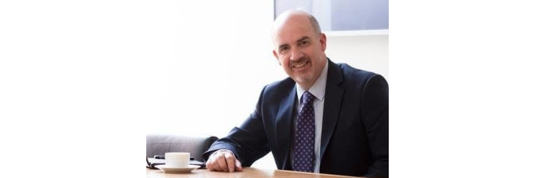 Professor Thom Brooks sitting at a desk, wearing a suit and tie. He is smiling at the camera