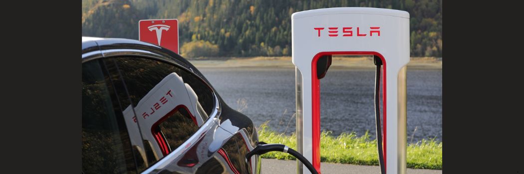 A close up of the rear wing of a black Tesla car as it charges at a red and white Tesla branded charging station.