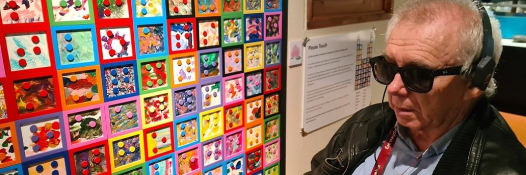 Man wearing sunglasses and headphones sits in front of a museum art exhibit.