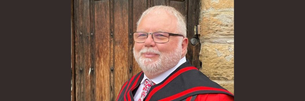 Professor Jon Gluyas with white hair, beard and glasses stands in front of an old wooden door in red and black academic robes.