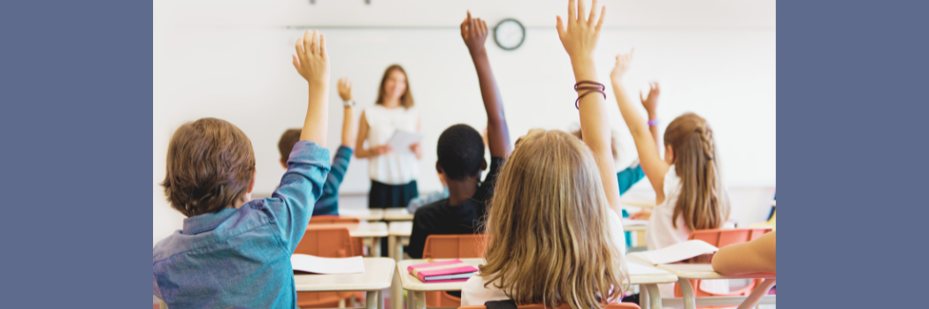 A teacher stands in front of a white board while students sitting in front of her raise their hands