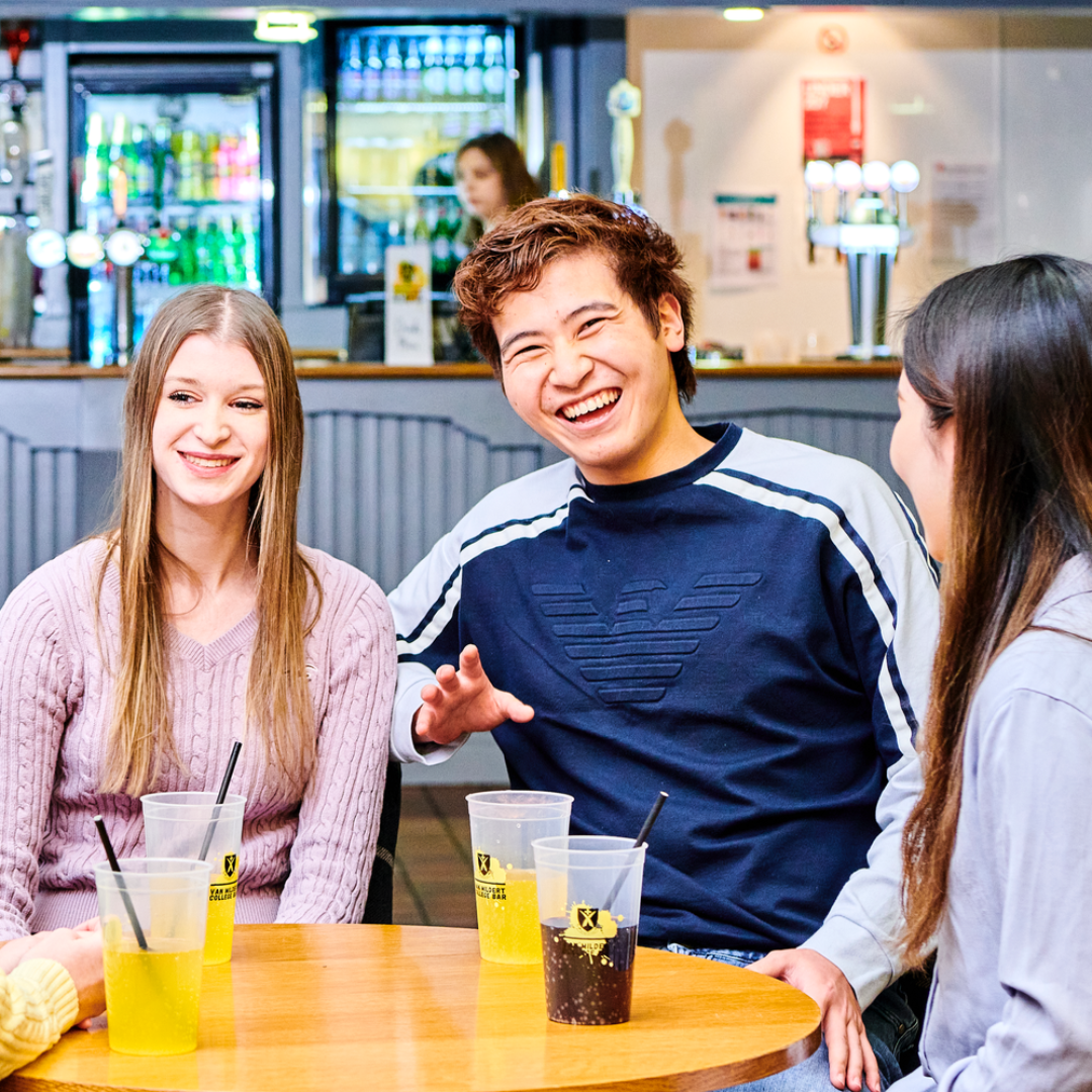 A group of students having a drink while seated around a table while several other students sit and talk at another table