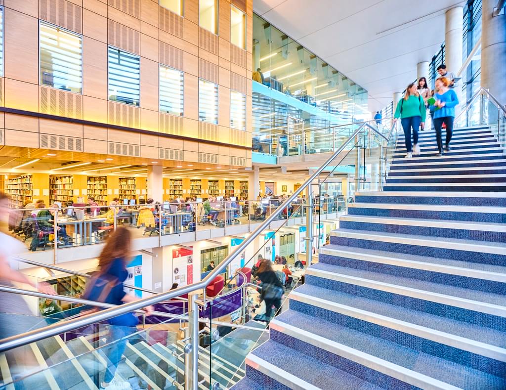 Timelapse photo of students walking up and down staircases inside a bright, airy and large university library.