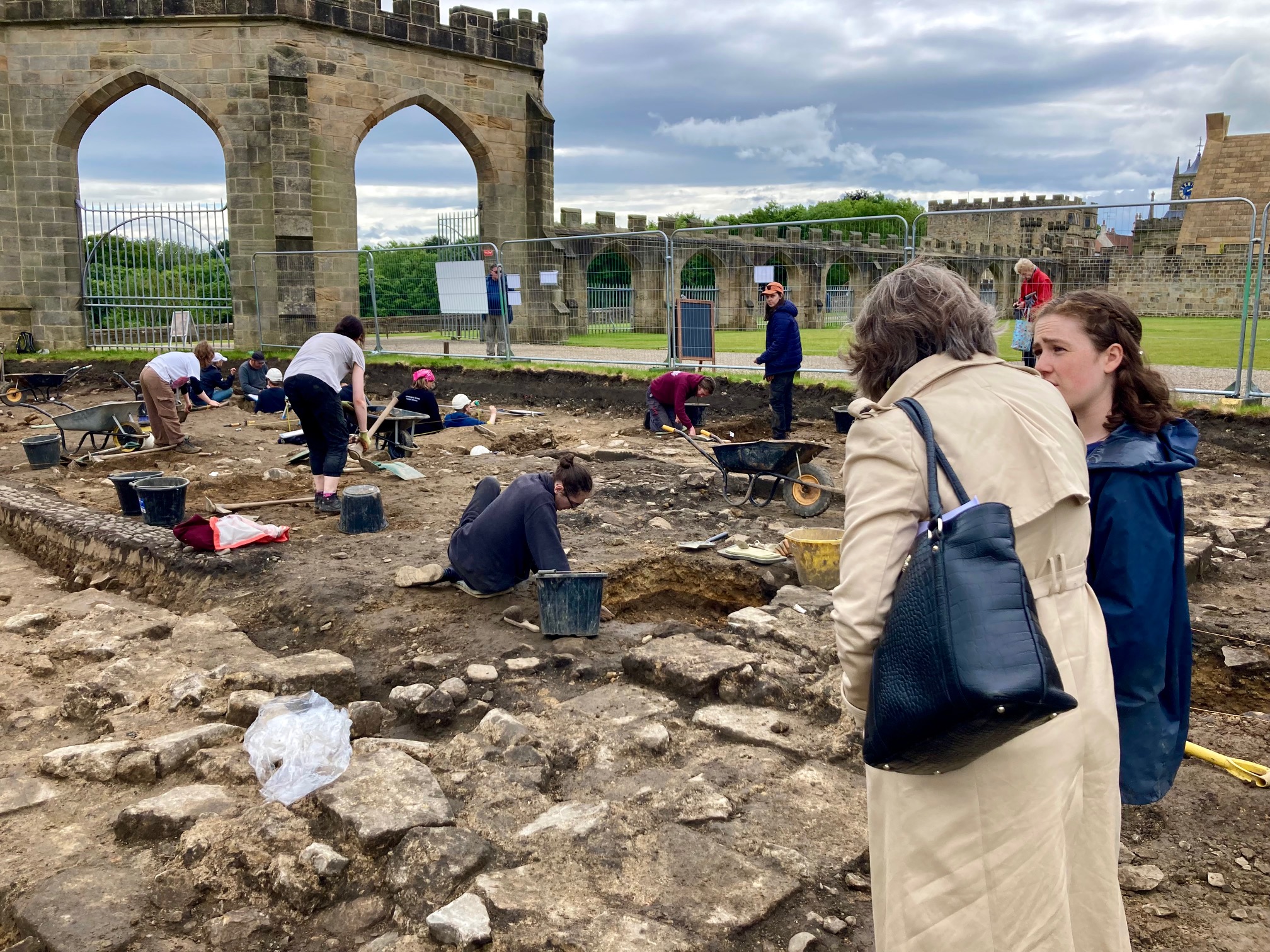 Two people are talking inside an archaeological trench, where remains of stone floors are gradually being uncovered. In the background are some Archaeology students with mattocks, buckets and shovels. In the far distance are arched medieval stone walls under a bluish grey cloudy sky.