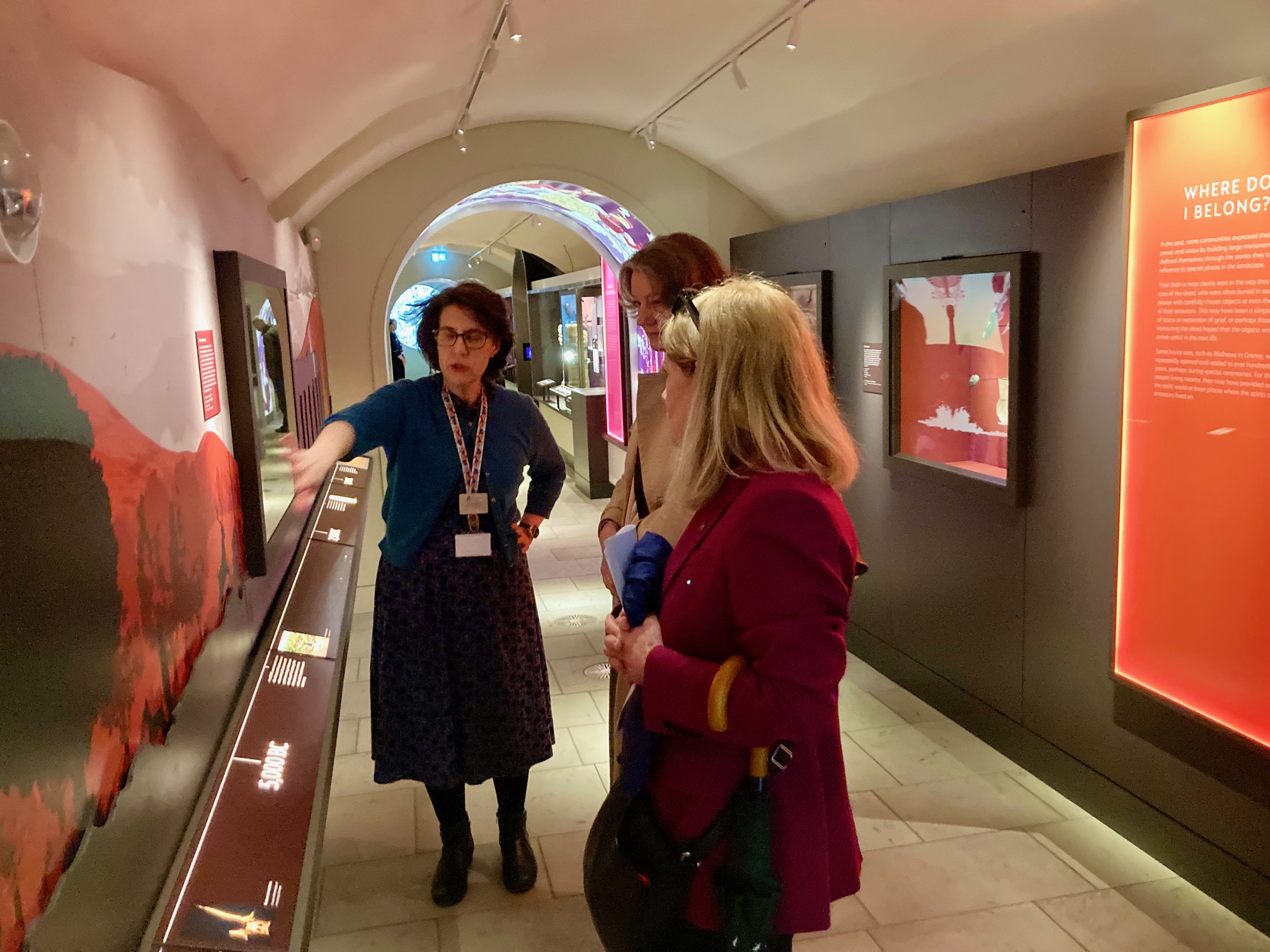 Three people stand in a warmly-lit museum corridor with glass cabinets displaying archaeological artefacts. One of the three is explaining a timeline on the wall to the other two people. The date 5000BC is visible.