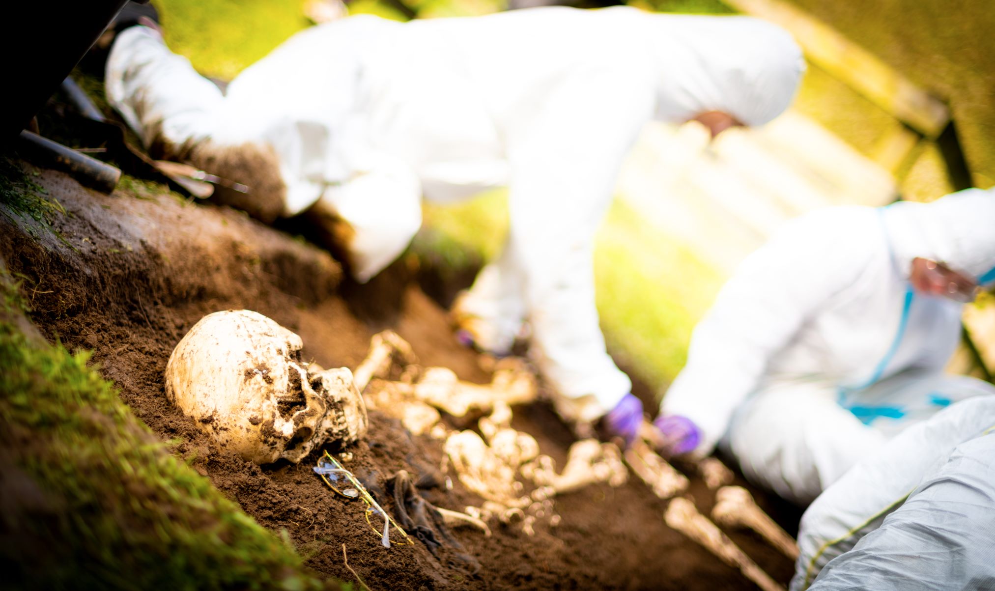 Angled photo of three people in white PPE excavating a simulated mass grave. In the foreground, in a shallow trench, are casts of some skeletonised human remains next to a pair of glasses and fabric scraps.