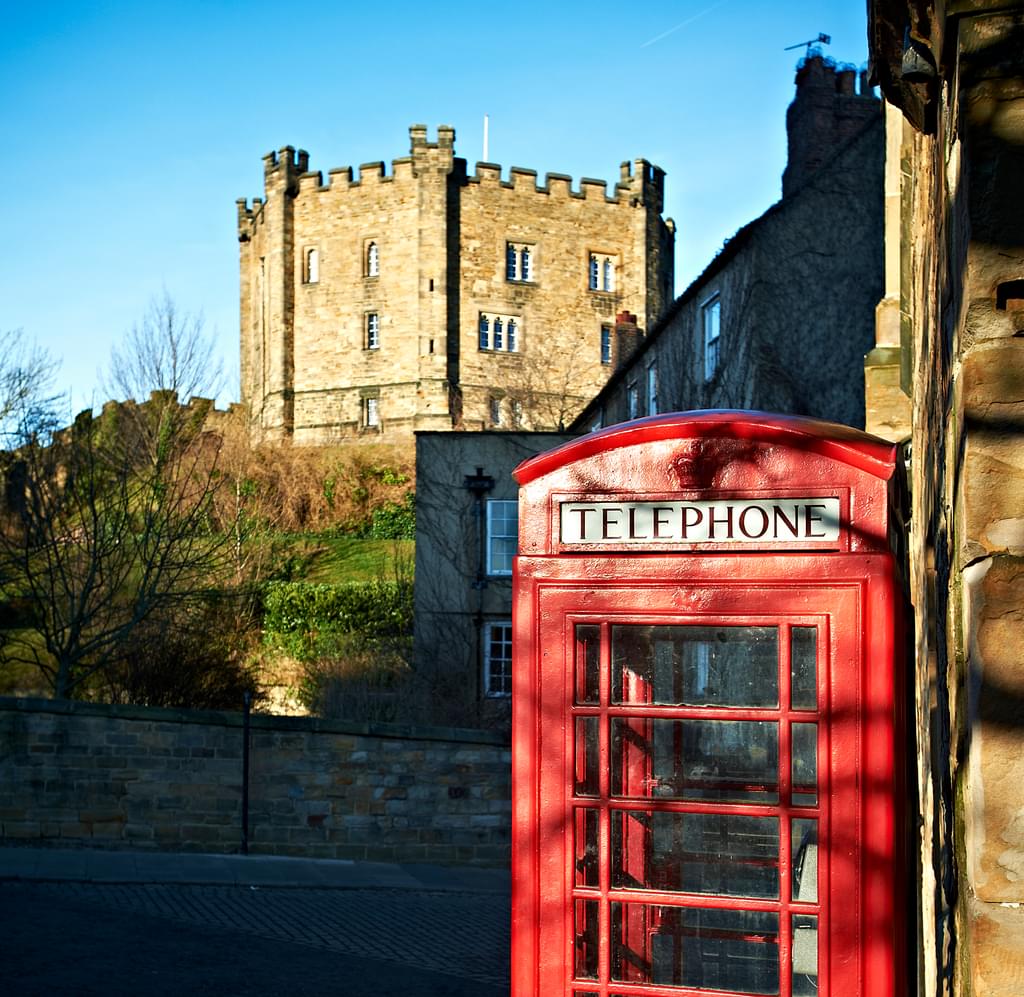 Golden hour light illuminates a red telephone box with Durham Castle and a blue sky in the background.