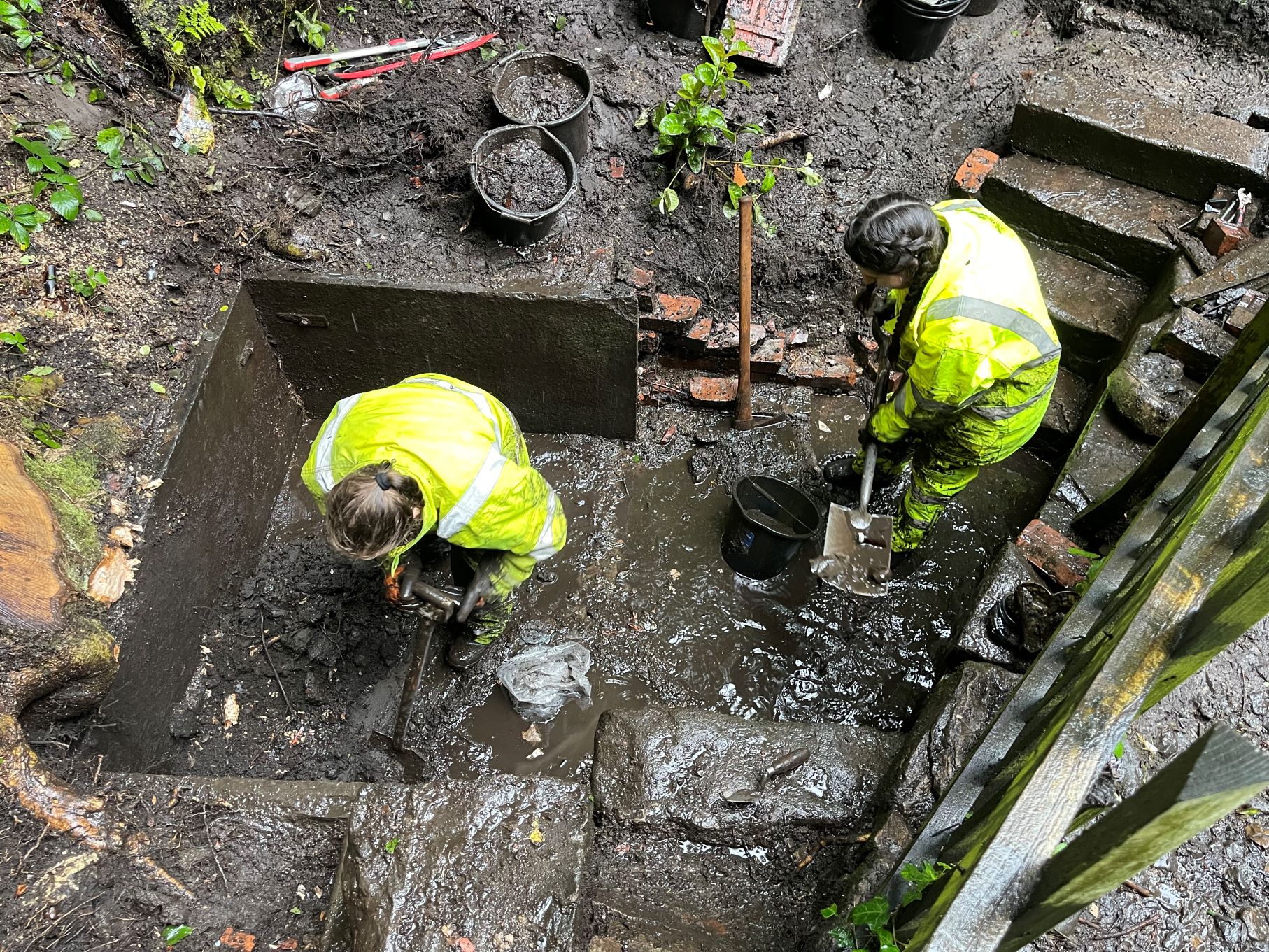 High angle photo of two archaeologists with high-vis jackets, waterproof attire, buckets and shovels standing in a wet and muddy trench next to some steps.