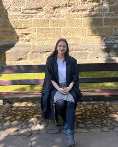 Student sitting on a low level bench by a stone wall outside Durham Cathedral wearing a University robe