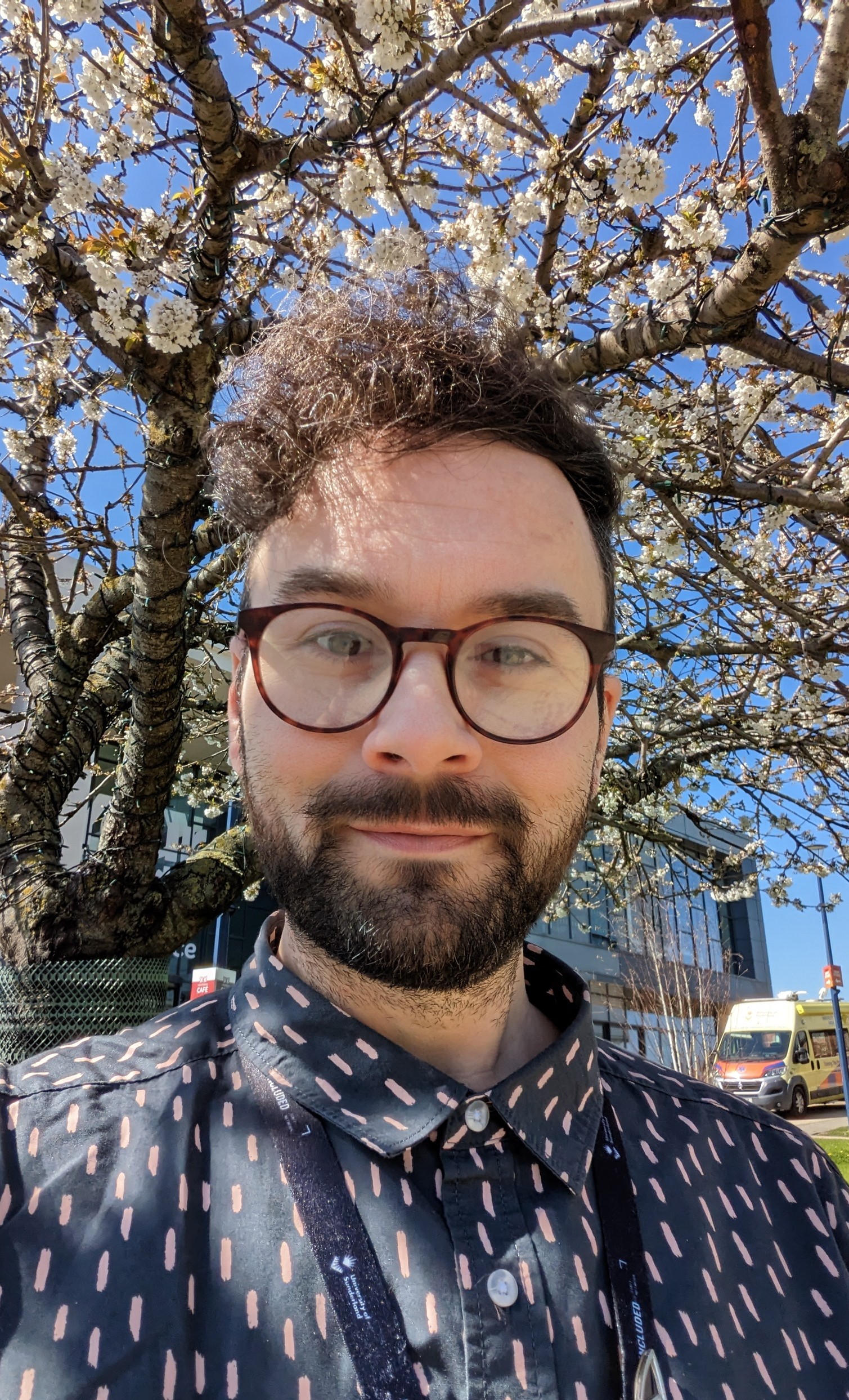 Andrew is light skinned with brown, curly hair and a brown beard. Andrew is wearing glasses and a navy and pink shirt. In the background are blue skies and a blossom tree