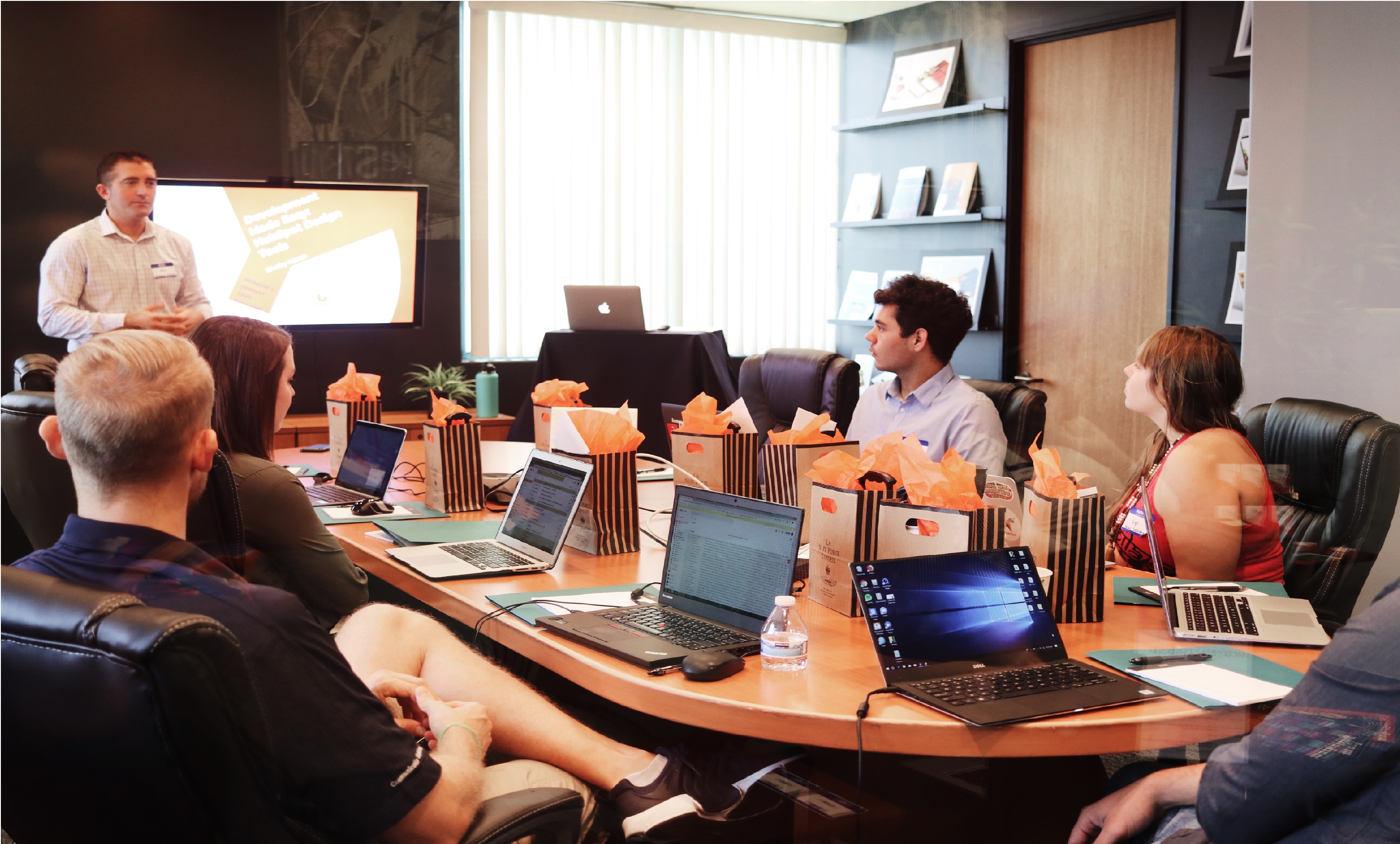 A meeting of people around a long desk, all with laptops out, facing someone giving a presentation.