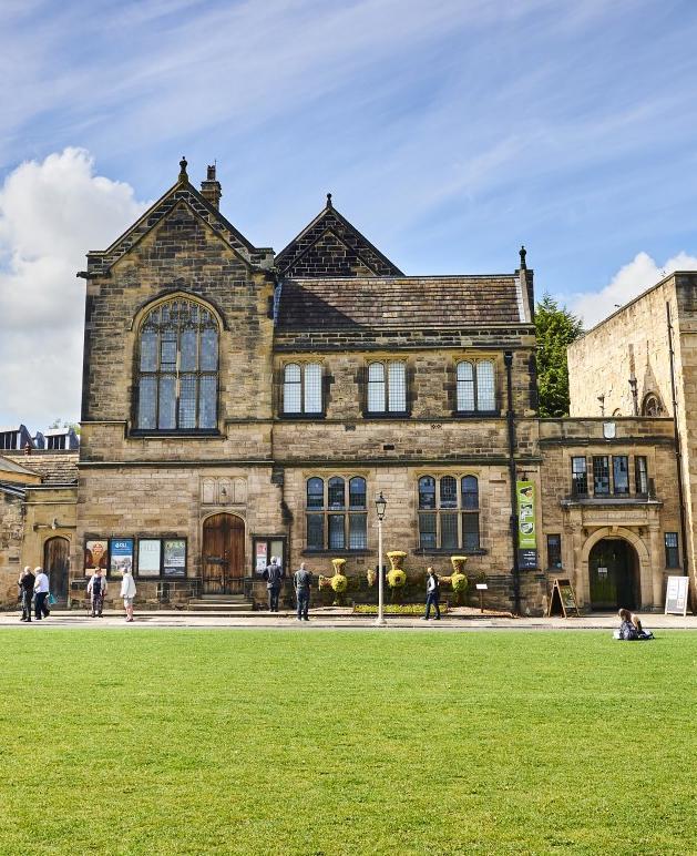 Series of historic stone buildings of different shapes and sizes sit behind a road way. There is a large lawn in the front.